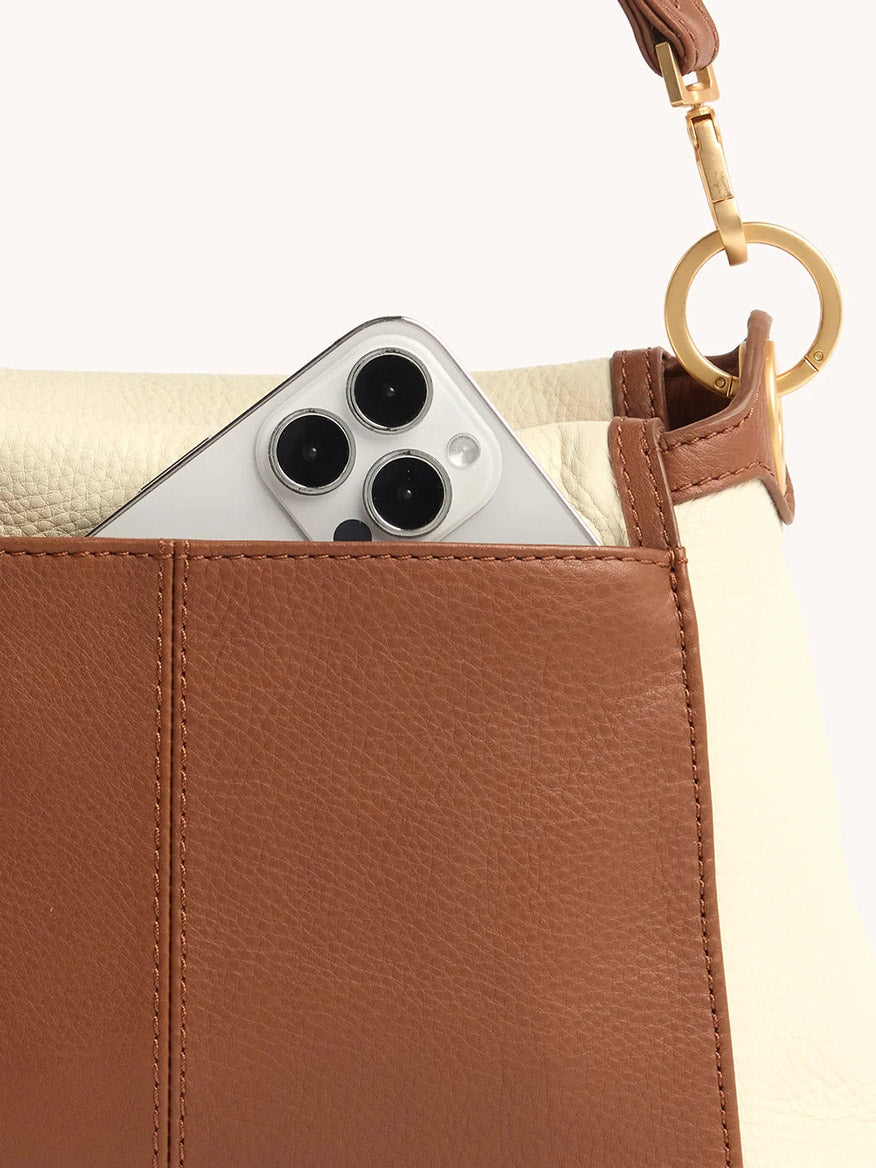 A smartphone with triple rear cameras peeks out from the front pocket of the Hammitt Los Angeles Bryant Medium Crossbody Bag in Courtyard Cream, which features brown and cream pebbled leather and a gold ring detail on the strap.