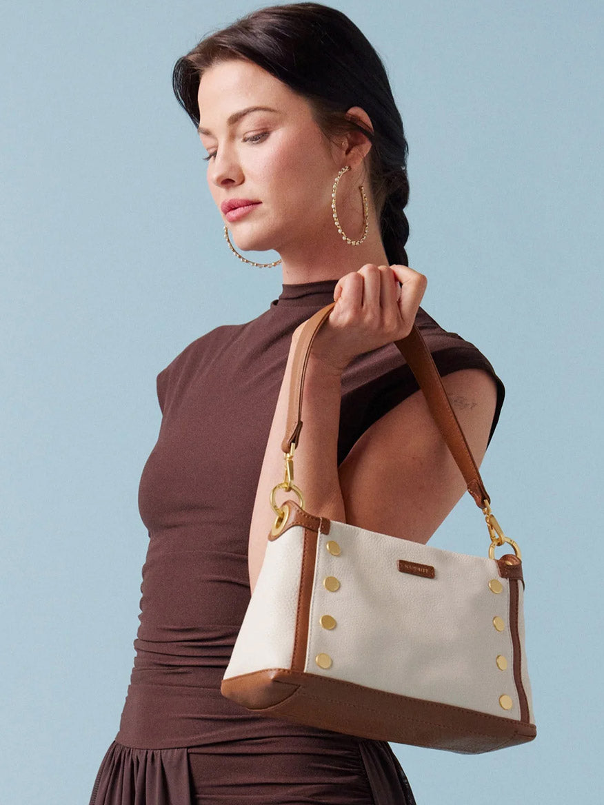 Woman in a brown dress holds the Hammitt Los Angeles Bryant Medium Crossbody Bag in Courtyard Cream, featuring white and brown pebbled leather with gold accents, against a plain light blue background.