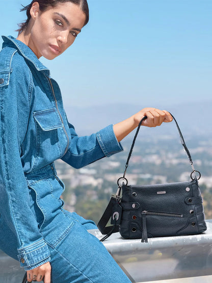 A person in a blue denim jumpsuit sits outdoors, holding the Hammitt Los Angeles Bryant Medium Crossbody Bag in Moto Black/Gunmetal, accented with metal eyelets and zippers. A cityscape and blue sky are in the background.