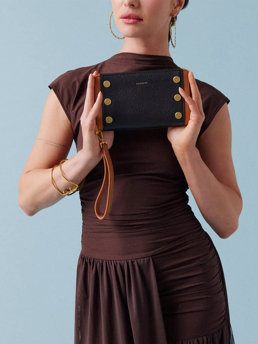 A woman in a brown dress holds the Hammitt Los Angeles Bryant Wallet in North End, made of soft black leather with gold studs and a detachable wristlet, against a light blue background.