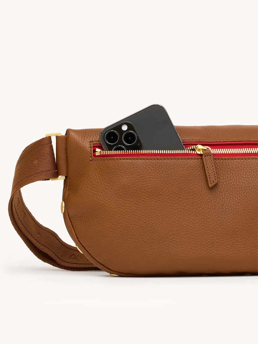 A black smartphone partially tucked into the red-zippered pocket of the Hammitt Los Angeles Charles Crossbody Large in Mahogany Pebble/Brushed Gold & Red Zip.