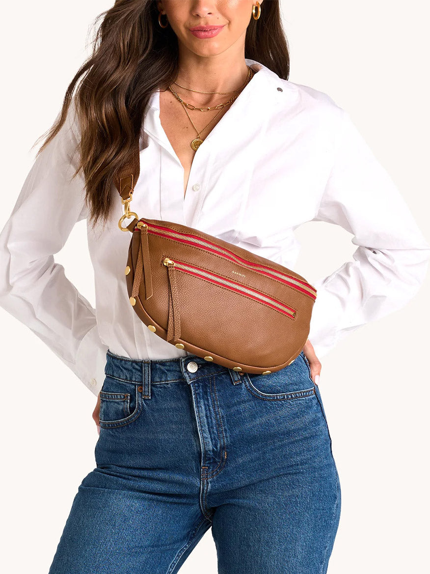 A woman in a white button-down and blue jeans poses with the Hammitt Los Angeles Charles Crossbody Large in Mahogany Pebble, featuring brushed gold studs and red zippers.