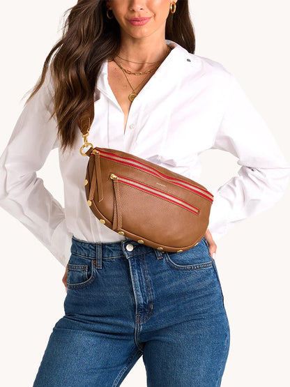 A woman in a white button-down and blue jeans poses with the Hammitt Los Angeles Charles Crossbody Large in Mahogany Pebble, featuring brushed gold studs and red zippers.