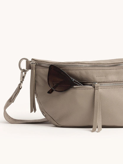 The Hammitt Los Angeles Charles Crossbody Large in Pewter Pebble/Brushed Silver is styled as a chic belt bag, with silver zippers and a pair of black sunglasses peeking from its front pocket.