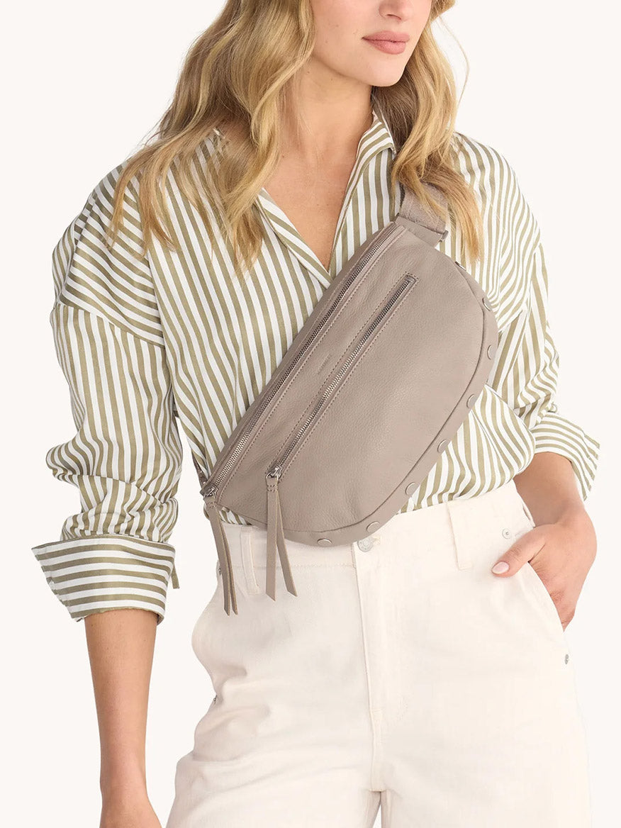 A woman in a beige striped shirt and white pants wears the Hammitt Los Angeles Charles Crossbody Large in Pewter Pebble/Brushed Silver slung across her chest.