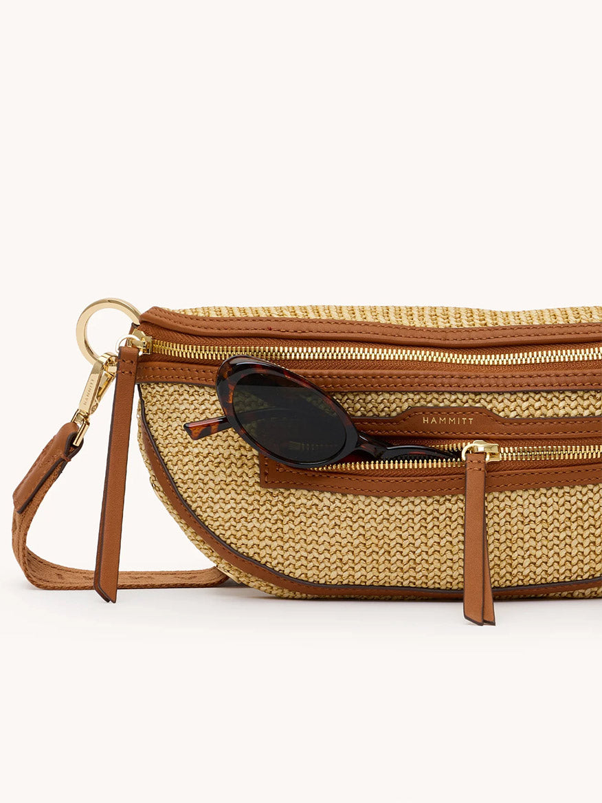The Hammitt Los Angeles Charles Crossbody Medium in Cocoa Raffia/Gold features woven straw, pebbled leather accents, gold hardware, zippered pockets, and a pair of sunglasses partially tucked in the front pocket.