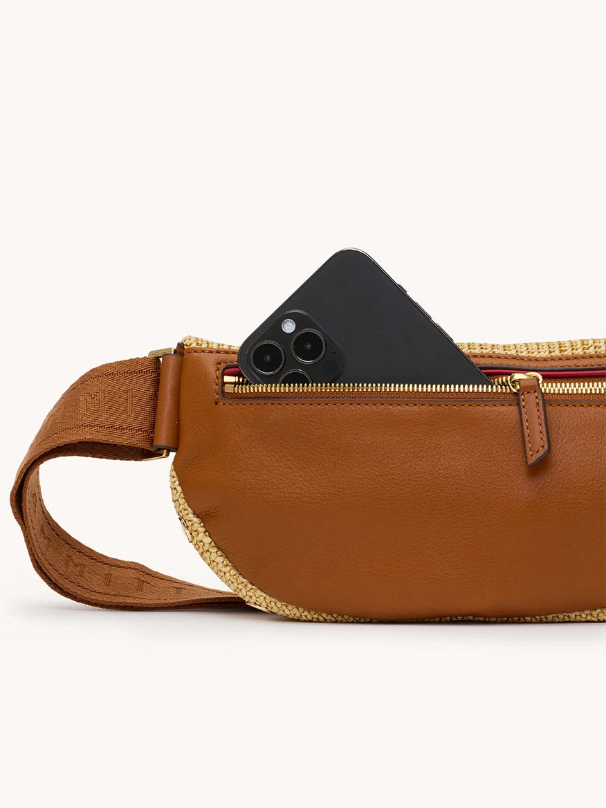 A black smartphone is partially tucked into the zippered pocket of the Hammitt Los Angeles Charles Crossbody Medium in Cocoa Raffia/Gold.