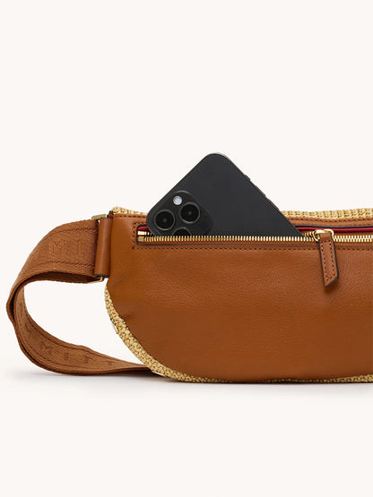 A black smartphone is partially tucked into the zippered pocket of the Hammitt Los Angeles Charles Crossbody Medium in Cocoa Raffia/Gold.
