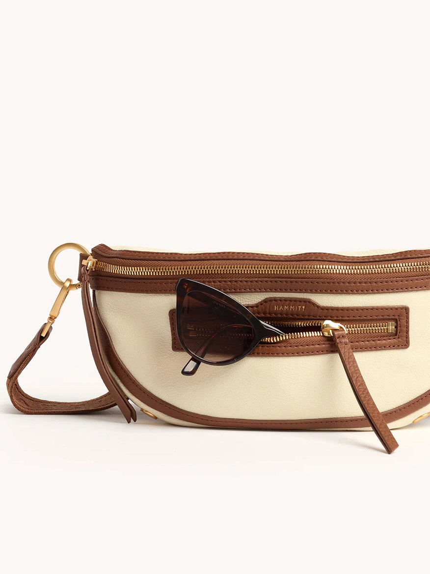 The Hammitt Los Angeles Charles Crossbody Medium in Courtyard Cream is a beige and brown pebbled leather bag with gold hardware, a front zipper pocket, and brown sunglasses hooked on the front.