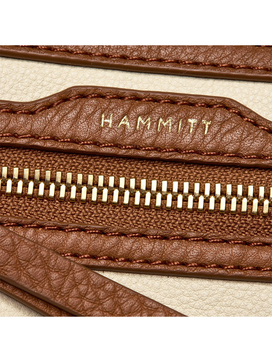 Close-up of the Hammitt Los Angeles Charles Crossbody Medium in Courtyard Cream, featuring a brown pebbled leather zipper with gold teeth and "HAMMITT" embossed in gold, set against a beige background.
