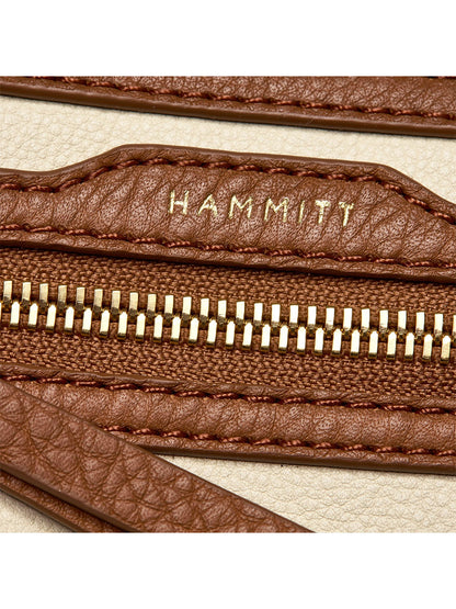 Close-up of the Hammitt Los Angeles Charles Crossbody Medium in Courtyard Cream, featuring a brown pebbled leather zipper with gold teeth and "HAMMITT" embossed in gold, set against a beige background.