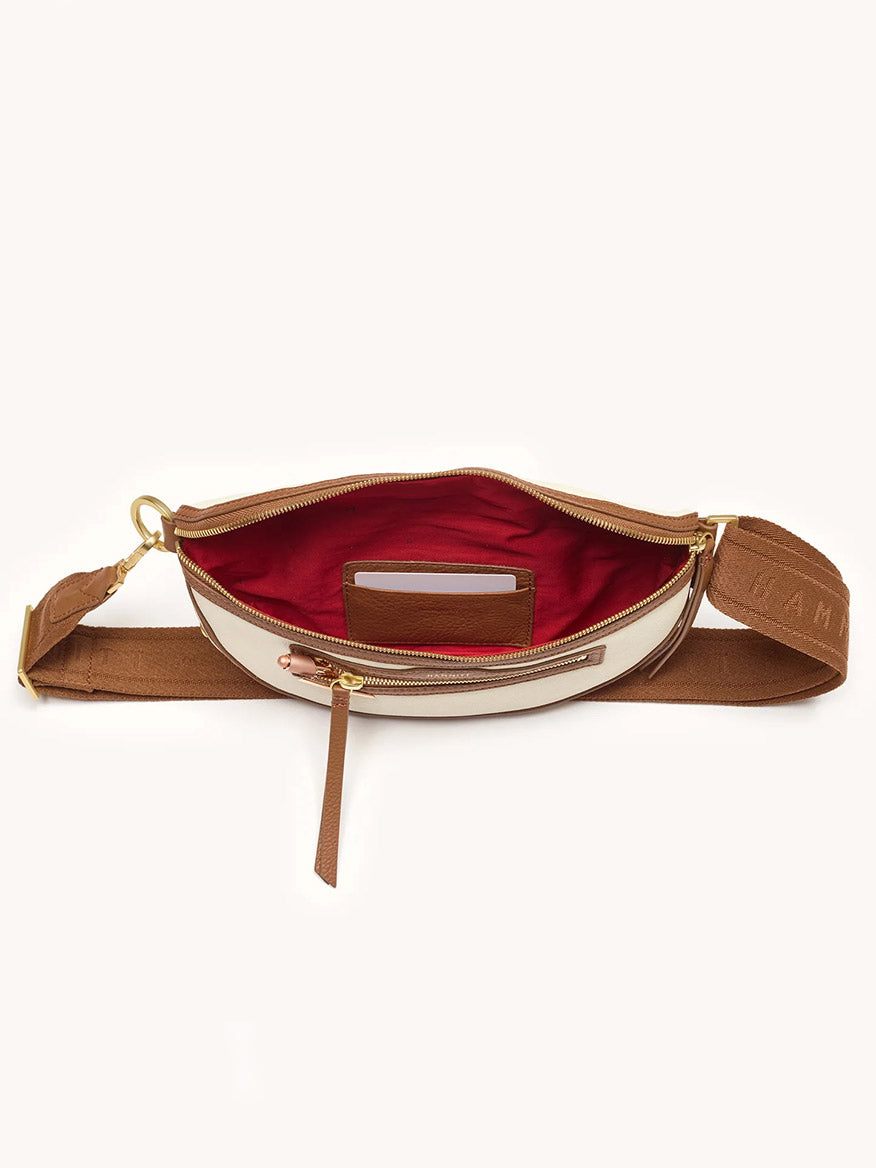 The Hammitt Los Angeles Charles Crossbody Medium in Courtyard Cream, featuring brown and white pebbled leather and a gold zipper, is open to display its red lining with a phone partially tucked into the inner pocket.
