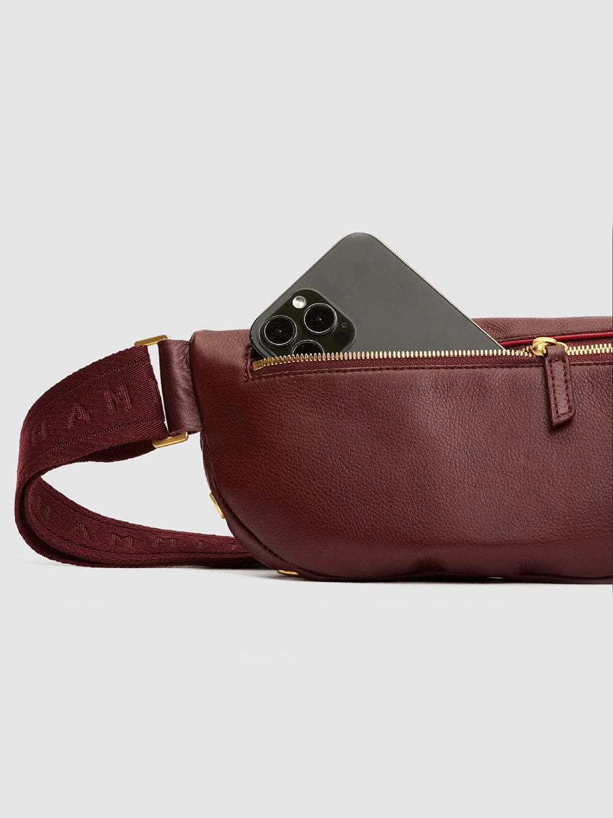 A black smartphone partially sticks out of the unzipped pocket of the Hammitt Los Angeles Charles Crossbody Medium in Cherrywood Pebble/Brushed Gold, a maroon pebble leather bag with a matching strap and gold zipper.