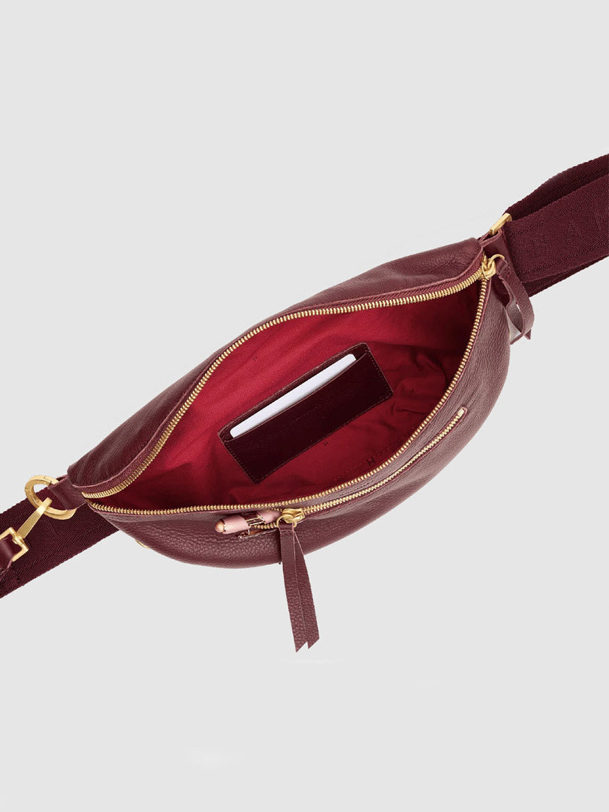 The Hammitt Los Angeles Charles Crossbody Medium in Cherrywood Pebble/Brushed Gold features a burgundy pebble leather exterior with a brushed gold zipper, shown open to reveal its vibrant red interior and a small card tucked into the inside pocket.