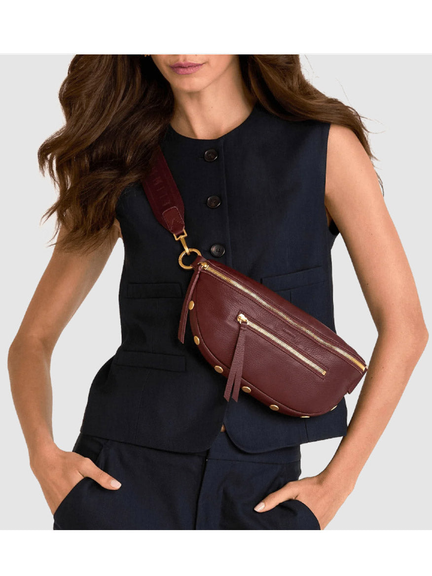 A woman in a sleeveless black buttoned vest and matching pants models the Hammitt Los Angeles Charles Crossbody Medium in Cherrywood Pebble/Brushed Gold—crafted from luxe pebble leather with brushed gold zippers and hardware—against a light gray background.