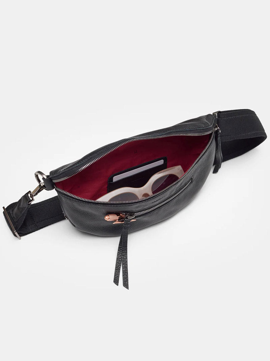 The Hammitt Los Angeles Charles Crossbody Medium in Black/Gunmetal features a stylish black pebbled leather exterior and a striking red interior. When unzipped, it unveils a pair of sunglasses and a card nestled inside.