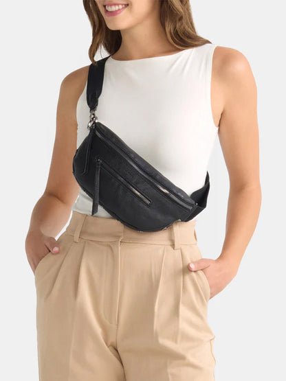 A woman is wearing a white sleeveless top and beige high-waisted pants, accessorized with the Hammitt Los Angeles Charles Crossbody Medium in Black/Gunmetal. She has her hands in her pockets.