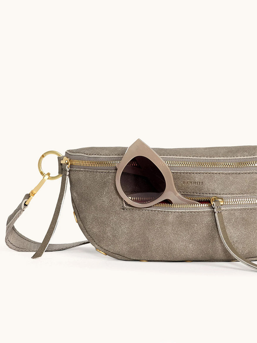 The Hammitt Los Angeles Charles Crossbody Medium in Pewter Brushed Nubuck/Brushed Gold, featuring gold hardware and a convenient zippered pocket, holds a pair of beige cat-eye sunglasses partially tucked inside.