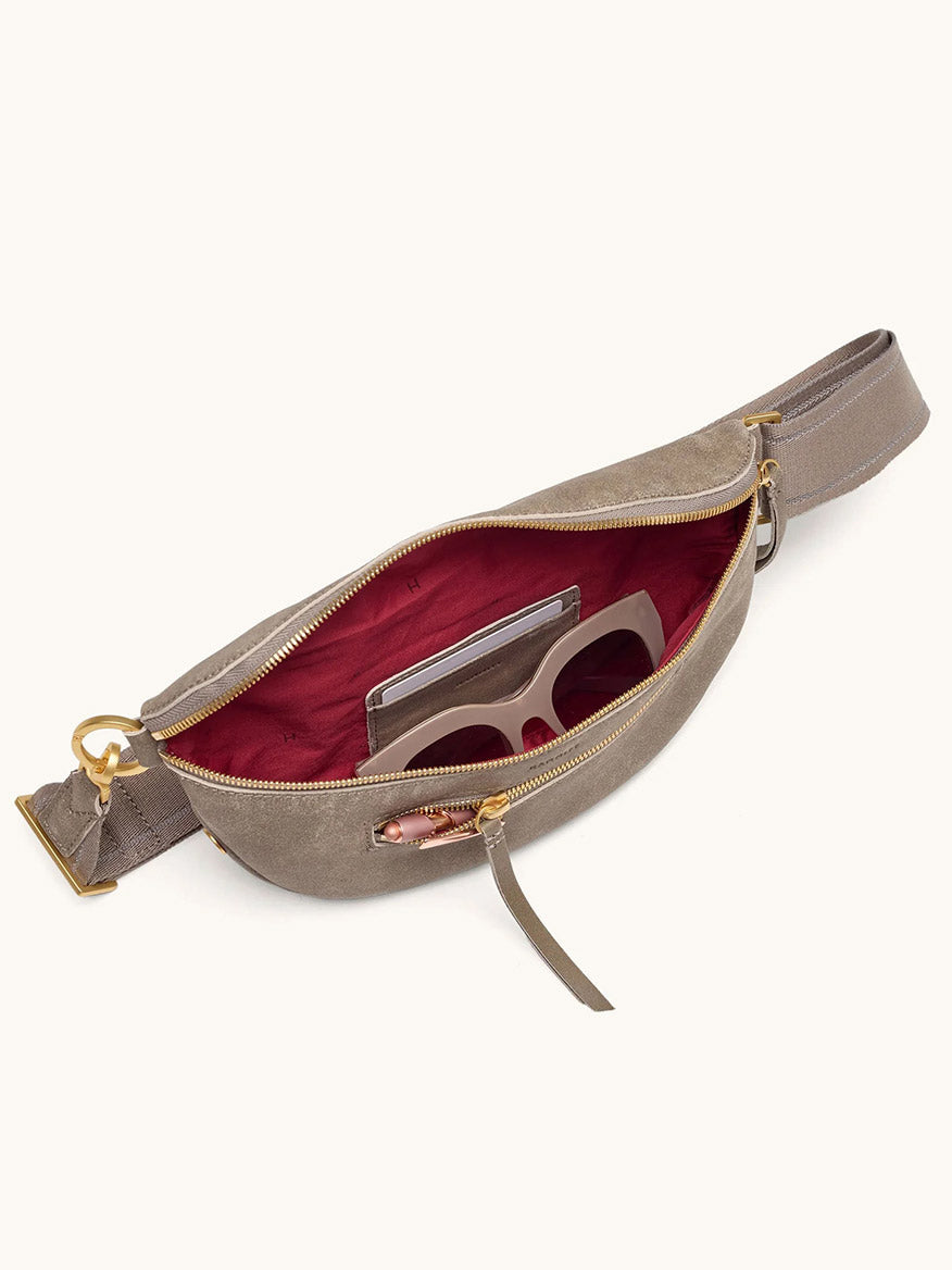 The Hammitt Los Angeles Charles Crossbody Medium in Pewter Brushed Nubuck/Brushed Gold features a chic pewter leather design with a brushed gold buckle and a striking red interior. Partially unzipped to reveal sunglasses and a smartphone inside, this versatile crossbody belt bag effortlessly combines style and practicality—perfect for carrying your essentials wherever you go.