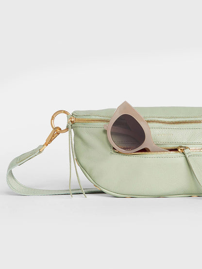 A Hammitt Los Angeles Charles Crossbody Medium in Cypress Sage with an open zipper revealing a pair of sunglasses partially sticking out, against a grey background.
