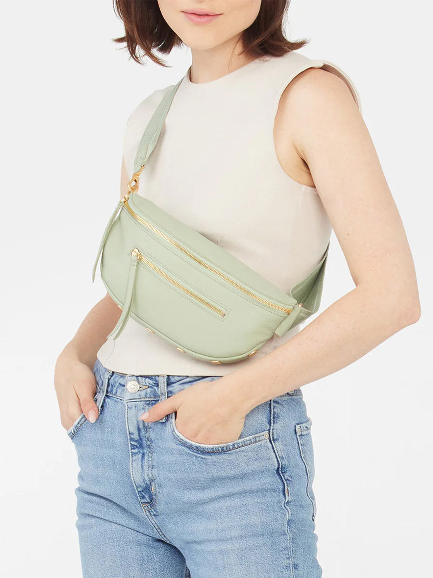 Woman in a white sleeveless top and blue jeans, wearing a Hammitt Los Angeles Charles Crossbody Medium in Cypress Sage with gold zippers.