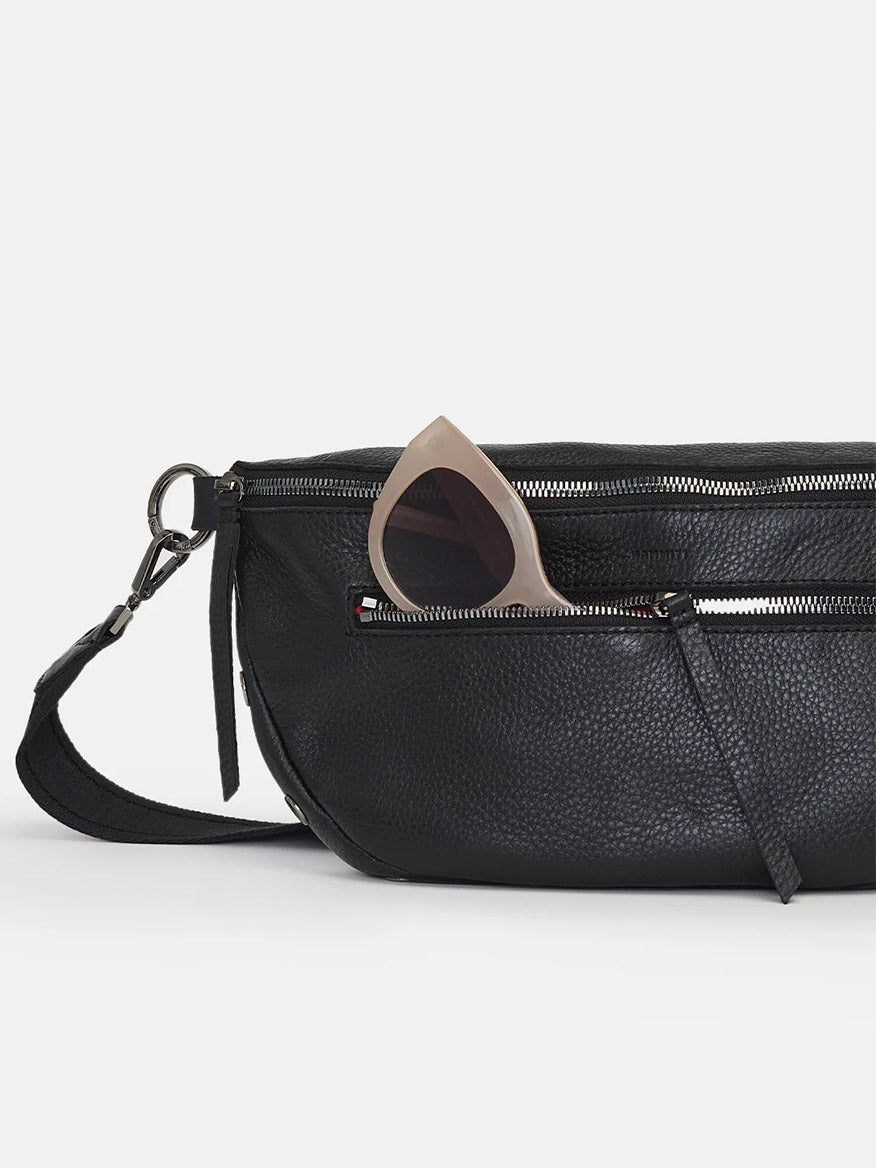 A Hammitt Los Angeles Charles Crossbody Large in Black with an adjustable crossbody strap and a pair of sunglasses partially tucked into the front zipper pocket, isolated on a white background.