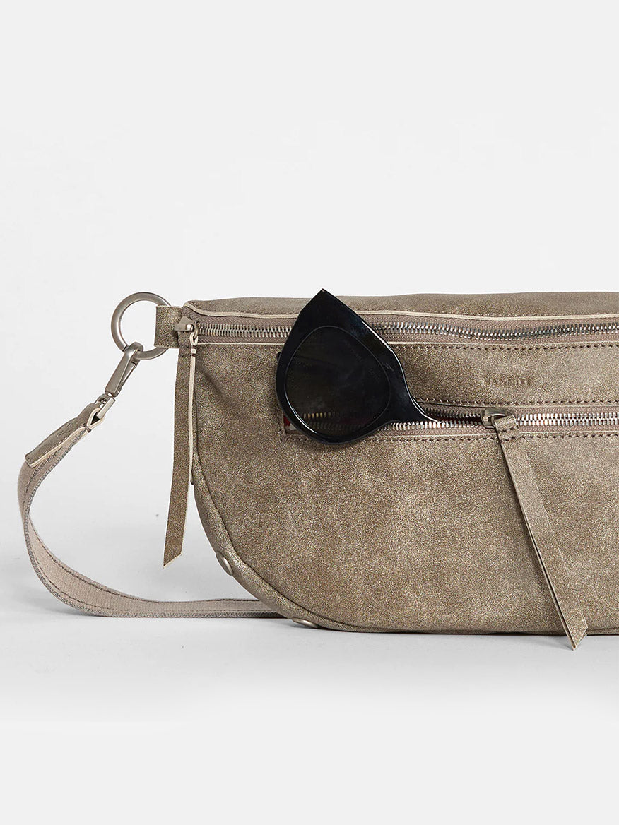 A Hammitt Los Angeles Charles Crossbody Large in Pewter with multiple zippers and leather zipper pulls, and a black sunglass resting on it against a plain background.