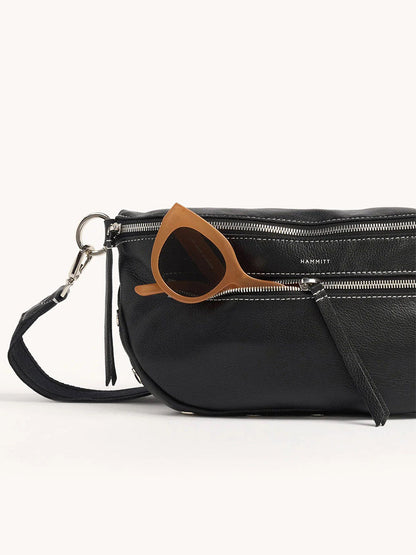 The Hammitt Los Angeles Charles Crossbody Large in Tempo Black is a chic black pebbled leather crossbody bag with an adjustable strap and multiple zippers, shown accessorized with brown sunglasses partially tucked into one compartment.