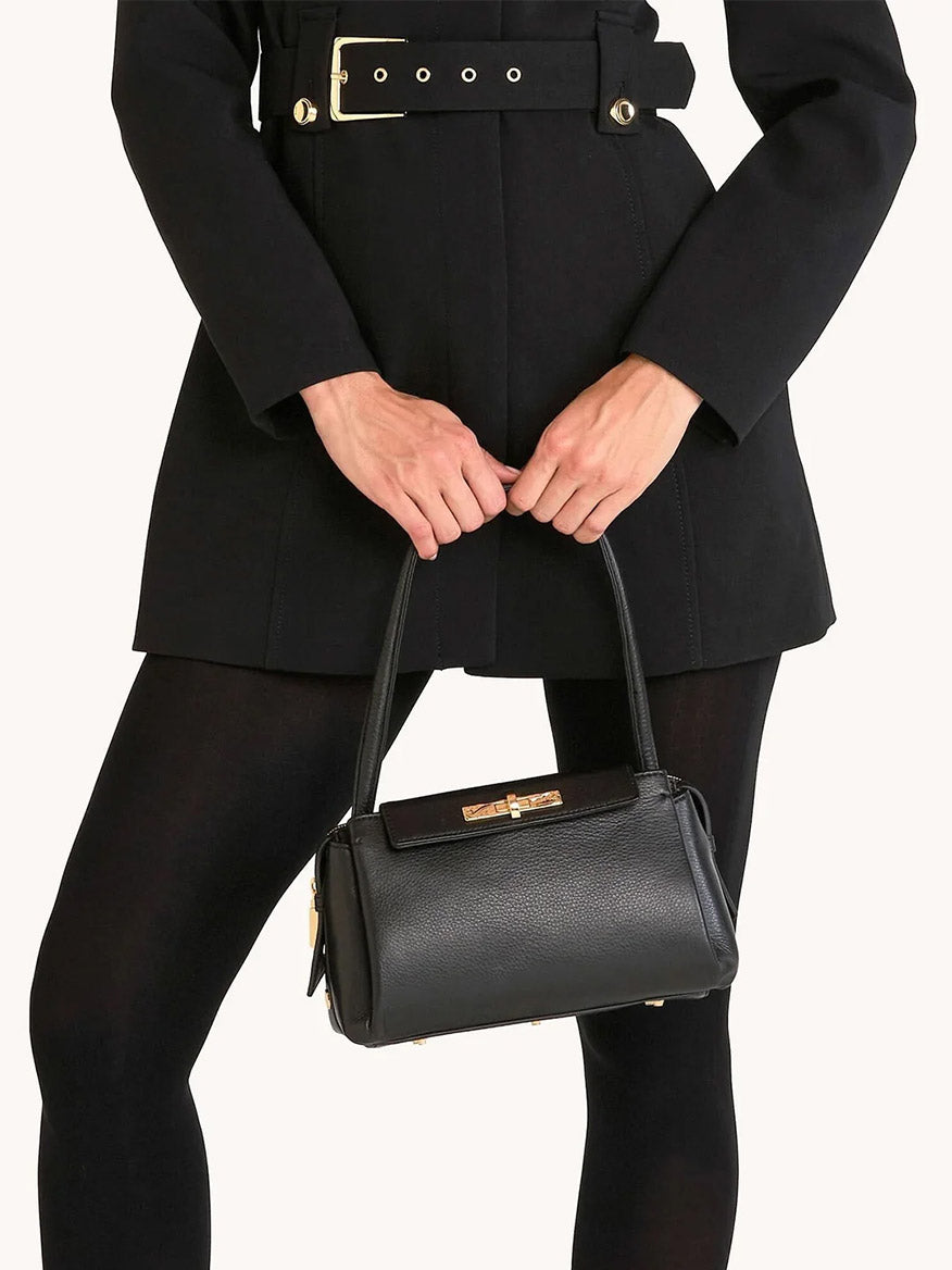 A person wearing a black belted coat and tights holds the Hammitt Los Angeles Charlotte Medium in Black/Gold, displaying its sleek pebble leather design, gold hardware, and spacious capacity in front of their body.