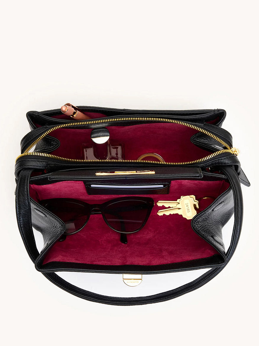 Open Hammitt Los Angeles Charlotte Medium in Black/Brushed Gold, with burgundy lining, shown from above holding sunglasses, keys, a small bottle, a tube, and a round compact—a roomy bag for keeping essentials organized and stylish.