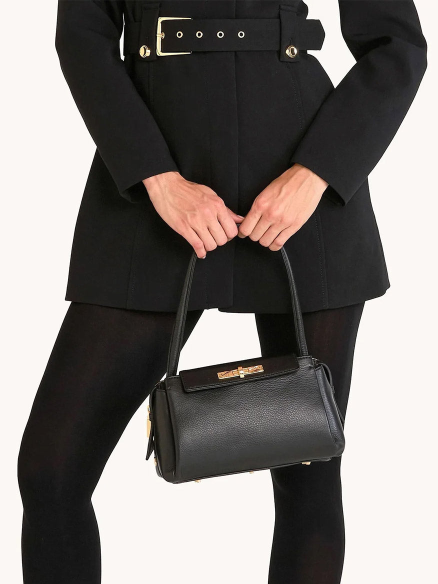 A person in a black belted coat and tights holds the Hammitt Los Angeles Charlotte Medium handbag in Black/Brushed Gold with gold hardware.