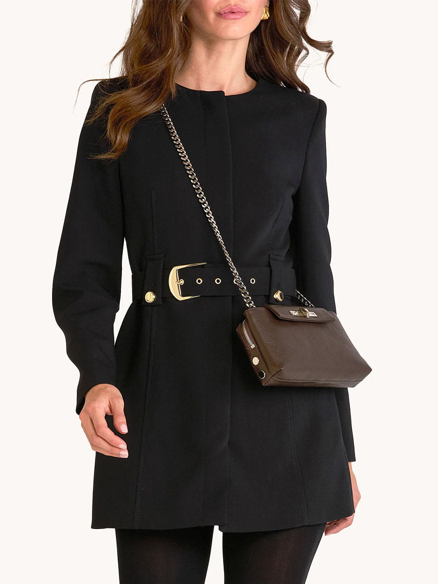 Woman in a tailored black coat with a belted waist holds the Hammitt Los Angeles Charlotte Small in Espresso Pebble/Silver crossbody with chain strap, paired with black leggings.