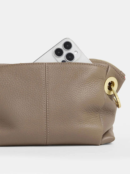 A smartphone with a triple-lens camera peeks out of the Hammitt Los Angeles Daniel Crossbody Clutch Small in Sculpted Taupe, featuring a gold grommet detail.