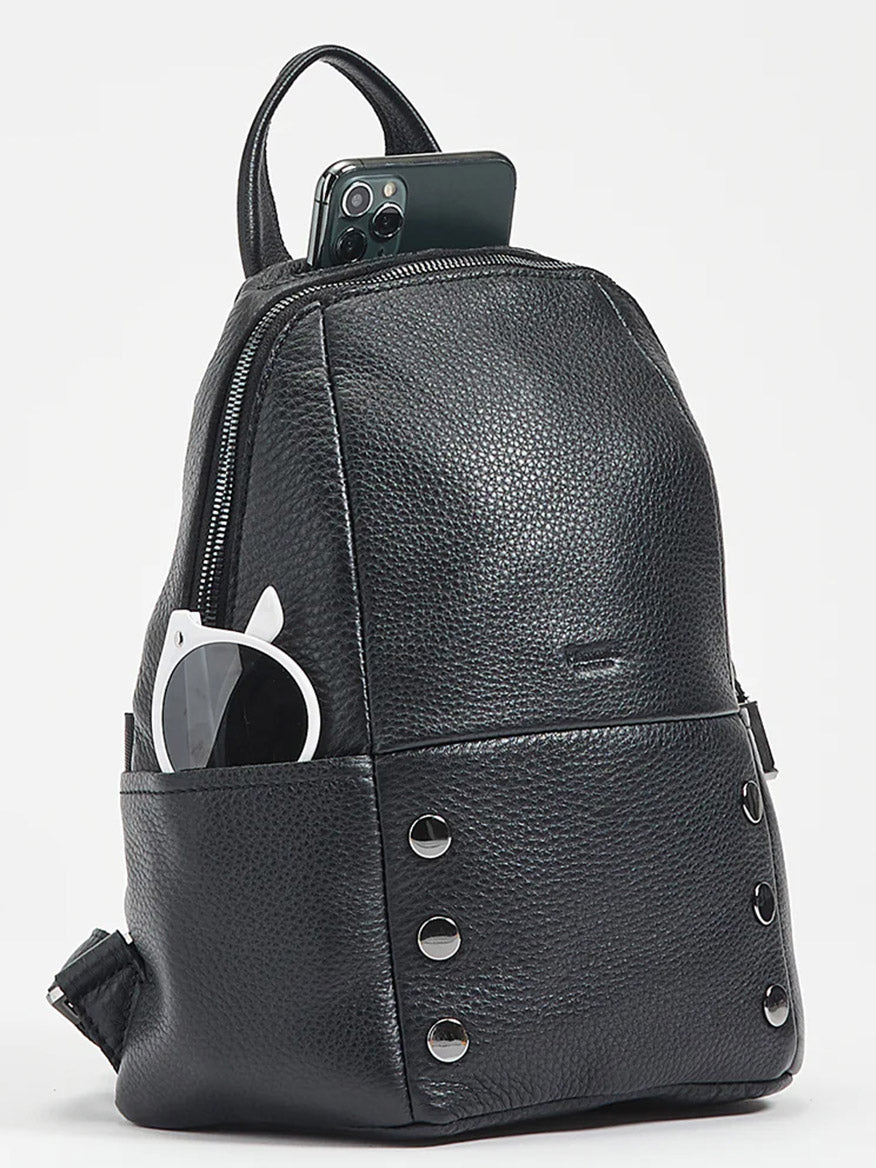 The Hammitt Los Angeles Hunter Medium Backpack in Black/Gunmetal, featuring silver studs and adjustable straps, is shown holding white sunglasses and a smartphone in its outer pockets against a plain background.