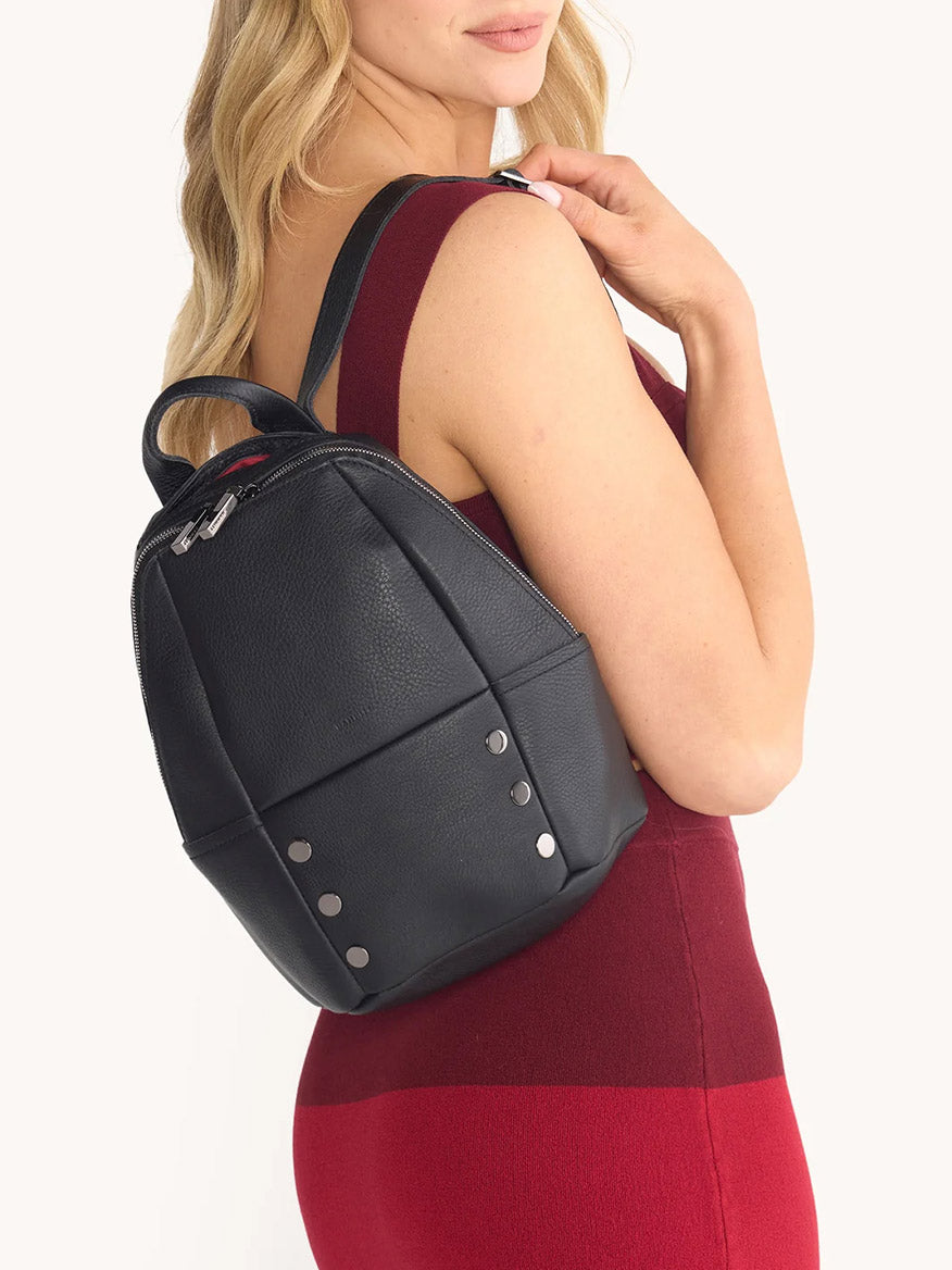 A woman in a sleeveless red dress carries the Hammitt Los Angeles Hunter Medium Backpack in Black/Gunmetal, featuring pebbled leather, silver studs, and adjustable straps over her shoulder.