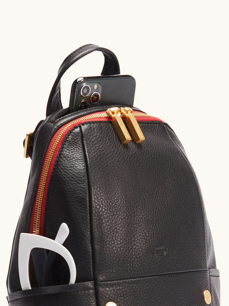 The Hammitt Los Angeles Hunter Medium Backpack in Black/Brushed Gold/Red Zip features a sleek design, with a smartphone in its top pocket and white sunglasses stored in the side pocket.