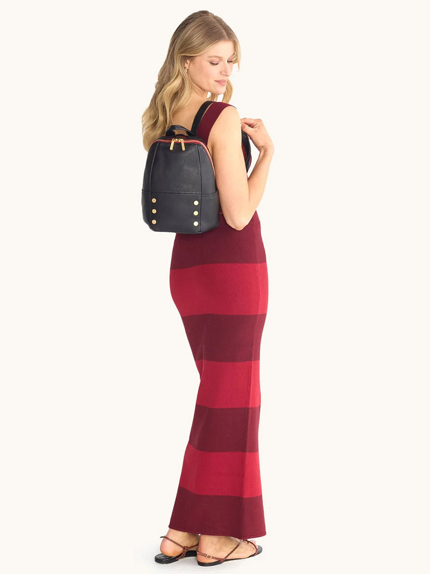 A woman with a sleeveless, red and burgundy striped maxi dress and sandals stands sideways, carrying the Hammitt Los Angeles Hunter Medium Backpack in Black/Brushed Gold/Red Zip.