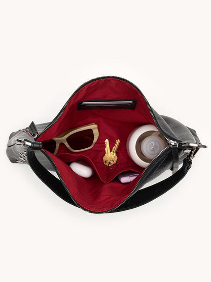 Open the Hammitt Los Angeles Kyle Large Shoulder Bag in Black/Gunmetal, revealing a red interior with sunglasses, a water bottle, lipstick, a compact mirror, and a keychain shaped like a hand making a peace sign inside.