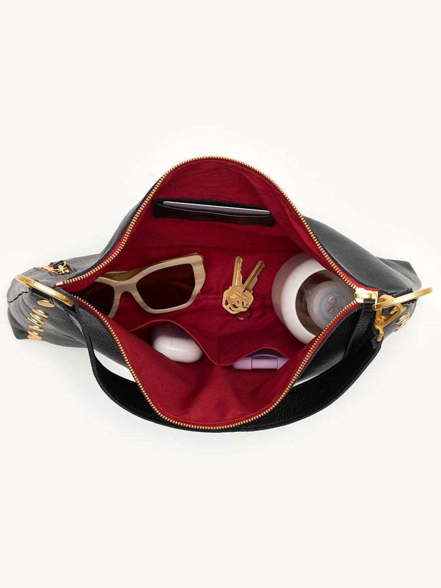 The Hammitt Los Angeles Kyle Large Shoulder Bag in Black/Brushed Gold & Red Zip displays sunglasses, a keychain, two round containers, and a bottle inside its spacious red-lined interior.
