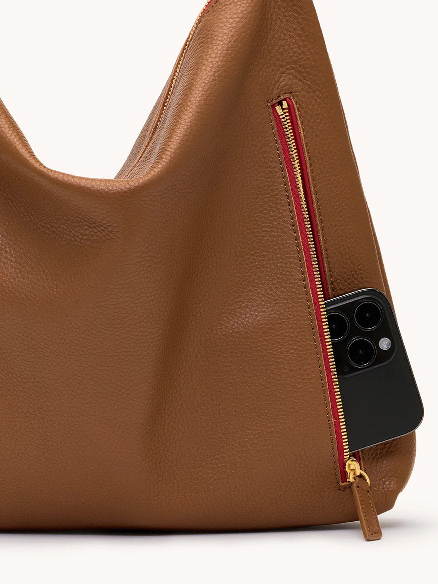A Hammitt Los Angeles Kyle Large Shoulder Bag in Mahogany Pebble/Brushed Gold/Red Zip has a side zipper pocket partially open, showing a smartphone inside.