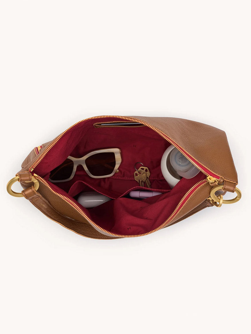 The Hammitt Los Angeles Kyle Large Shoulder Bag in Mahogany Pebble/Brushed Gold/Red Zip is open to reveal white sunglasses, keys, a white bottle, and a small container inside.