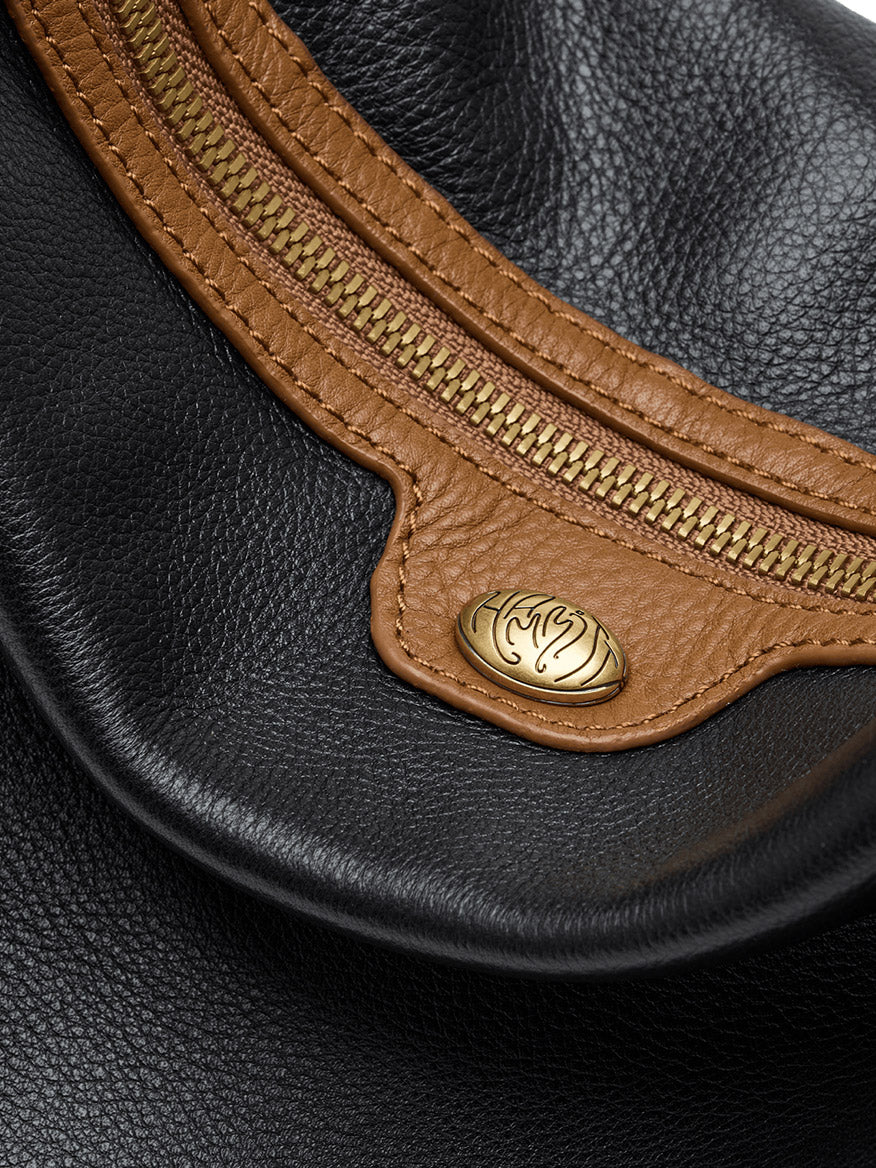 Close-up of a Hammitt Los Angeles Kyle Large Shoulder Bag in East End/Antique Gold, featuring black pebbled leather with brown trim, a gold zipper, and a round gold emblem with an abstract design.