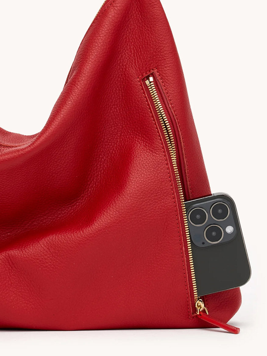 A black smartphone peeks from the zippered pocket of the Hammitt Los Angeles Kyle Large Shoulder Bag in Chili Red/Gold.