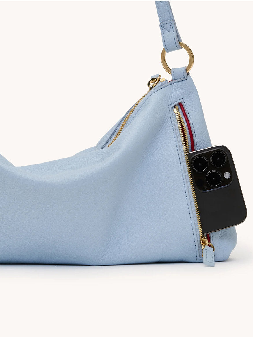 The Hammitt Los Angeles Kyle Medium Shoulder Bag in Hazy Blue/Gold is a pebbled leather crossbody with a gold zipper, exterior pocket holding a black smartphone, and is perfect for carrying daily essentials in style.