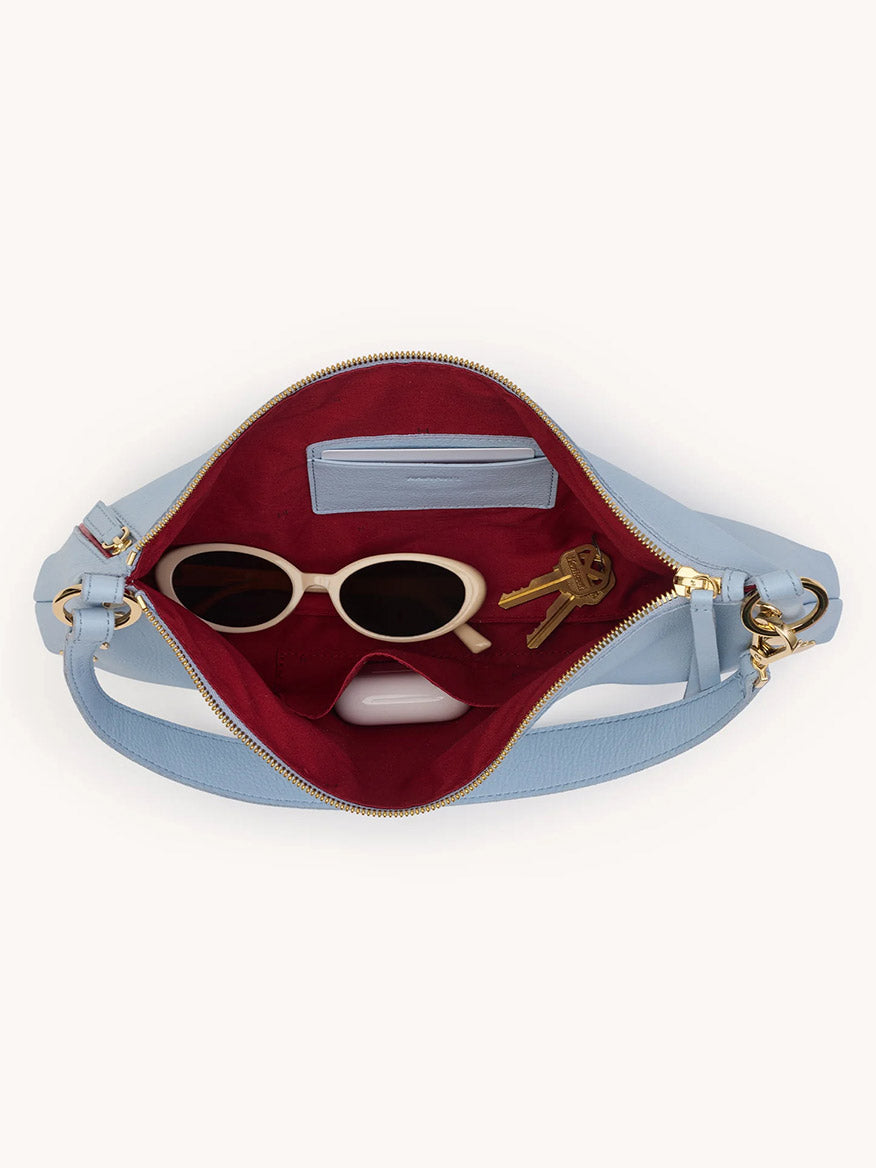 Open Hammitt Los Angeles Kyle Medium Shoulder Bag in Hazy Blue/Gold, revealing a red interior with white sunglasses, a blue cardholder, and keys inside.