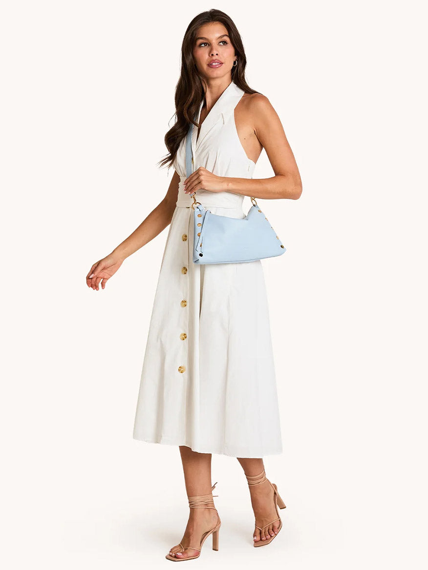 A woman in a sleeveless white midi dress with buttons and nude heels stands against a plain background, carrying the Hammitt Los Angeles Kyle Medium Shoulder Bag in Hazy Blue/Gold.