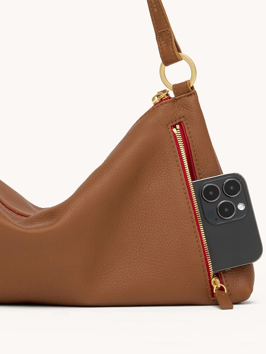 A medium brown Hammitt Los Angeles Kyle Medium Shoulder Bag in Mahogany Pebble with a brushed gold and red zipper, partially unzipped to reveal a black smartphone inside—a practical and stylish everyday accessory.