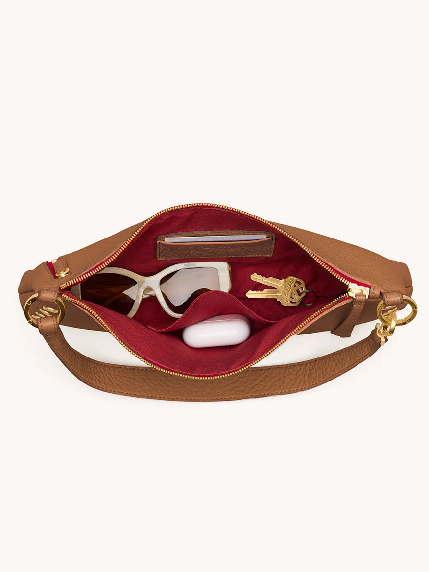 The Hammitt Los Angeles Kyle Medium Shoulder Bag in Mahogany Pebble/Brushed Gold & Red Zip is open, revealing sunglasses, a white case, keys, and a wallet organized inside its red interior.