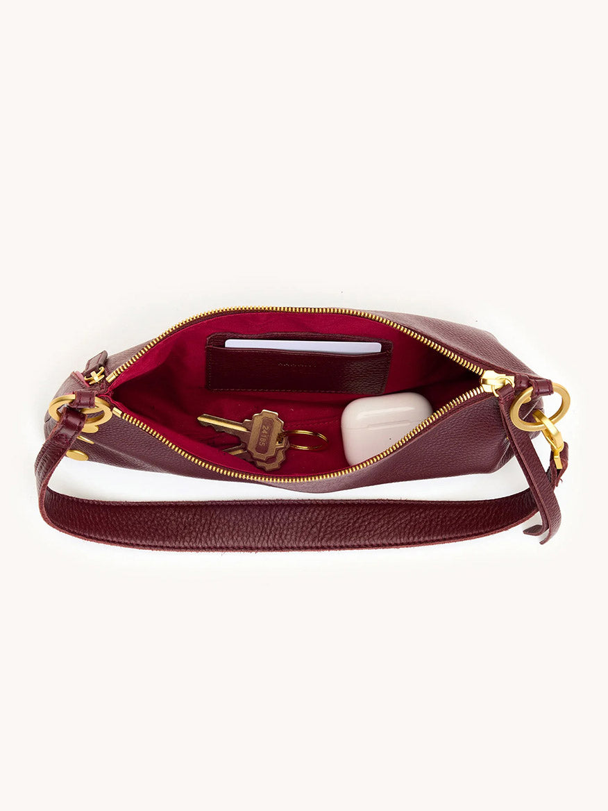 The Hammitt Los Angeles Kyle Shoulder Bag in Cherrywood Pebble/Brushed Gold features an adjustable crossbody strap, gold zipper, and red lining. Shown open with a phone, key, and white case inside.