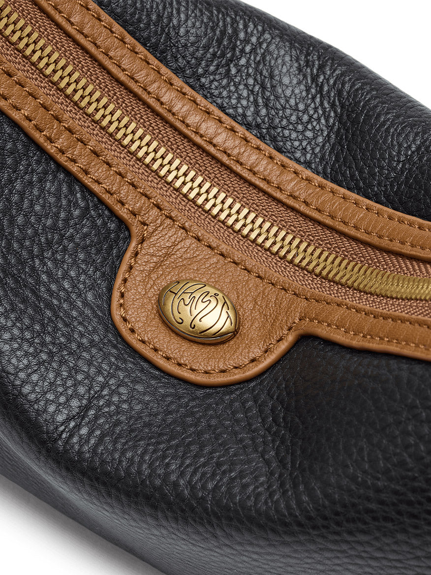 Close-up of the Hammitt Los Angeles Kyle Shoulder Bag in East End/Antique Gold, featuring black pebbled leather with brown leather trim, an antique gold zipper, and an oval gold emblem engraved with a decorative design.